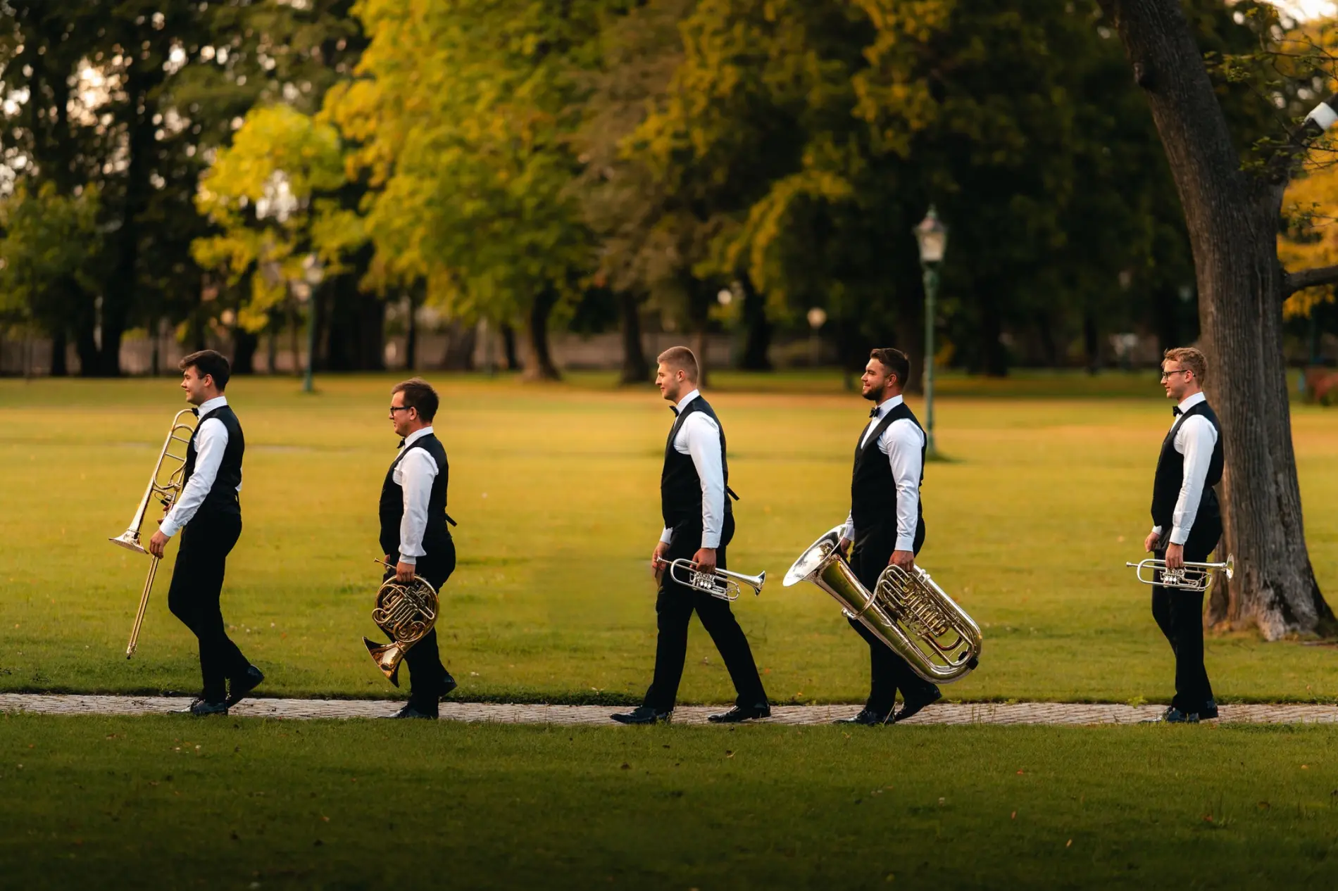 Muzycy Silver Brass w białych koszulach i kamizelkach idą przez park z instrumentami